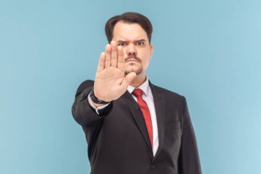 Portrait of strict bossy man with mustache showing block gesture, trying to stop conflict, looking at camera, wearing black suit with red tie. Indoor studio shot isolated on light blue background.
