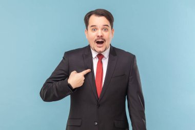 Portrait of shocked astonished man looking at camera with big eyes, pointing at himself, being surprised, wearing black suit with red tie. Indoor studio shot isolated on light blue background.
