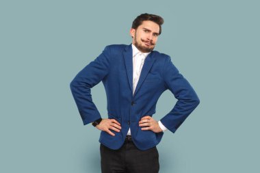 Portrait of funny angry man with mustache standing with hands on hips, looking at camera with frowning face, wearing official style suit. Indoor studio shot isolated on light blue background.