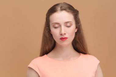 Closeup portrait of winsome adorable woman with long hair, wearing elegant dress, standing closed eyes, having calm expression. Indoor studio shot isolated on brown background.