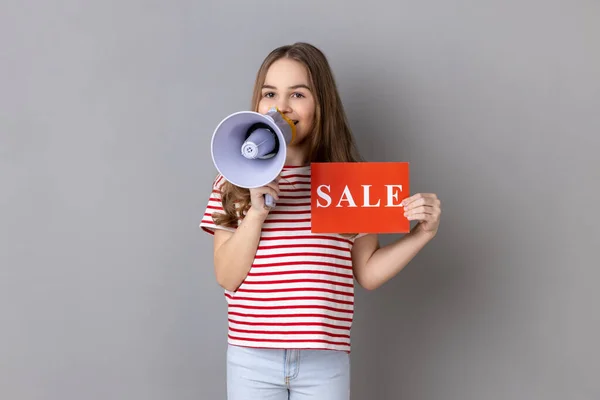 Little girl wearing striped T-shirt holding card with sale inscription and megaphone near mouth, loudly speaking, screaming, making announcement. Indoor studio shot isolated on gray background.