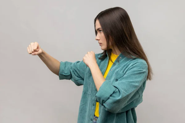 Side view of angry woman standing with clenched fists and furious look, ready to punch, expressing aggression, wearing casual style jacket. Indoor studio shot isolated on gray background.