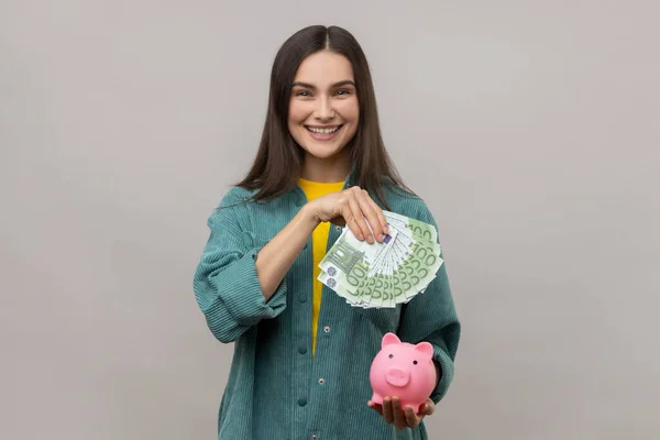 Positive attractive smiling woman holding euro banknotes and piggy bank, savings, high rate deposit, wearing casual style jacket. Indoor studio shot isolated on gray background.