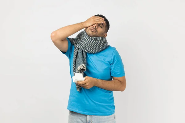 Portrait of sick ill unheatlhy unshaven man wearing blue T- shirt and wrapped in warm scarf standing touching forehead, has headache, has flu symptoms. Indoor studio shot isolated on gray background.