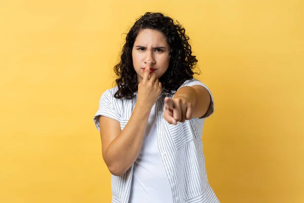I don\'t believe, you are liar. Dissatisfied woman with dark wavy hair in business suit touching nose showing lie gesture, suspecting cheating. Indoor studio shot isolated on yellow background.