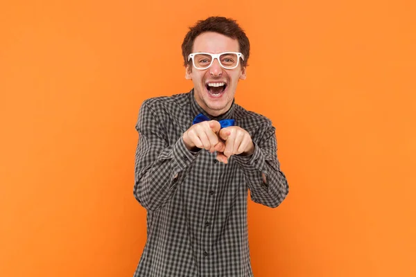 Overjoyed man nerd laughs at something funny, points directly into camera, expresses good emotions, makes choice, wears shirt with bow tie and glasses. Indoor studio shot isolated on orange background