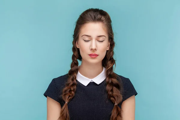 Portrait of attractive beautiful calm woman with braids standing with closed eyes, having problems with eyesight, wearing black dress. woman Indoor studio shot isolated on blue background.