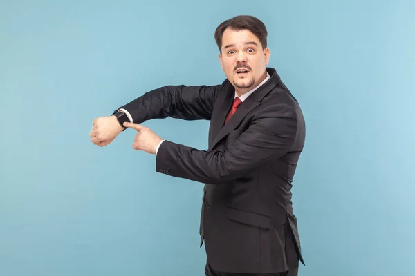 Confused man with mustache standing and looking at camera with puzzled expression, pointing at his wristwatch, wearing black suit with red tie. Indoor studio shot isolated on light blue background.