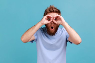 Şaşırmış sakallı adamın portresi elleriyle dürbün hareketi yapıyor ve kameraya hayret içinde bakıyor, ağzını açık tutuyor. Mavi arka planda kapalı stüdyo çekimi.