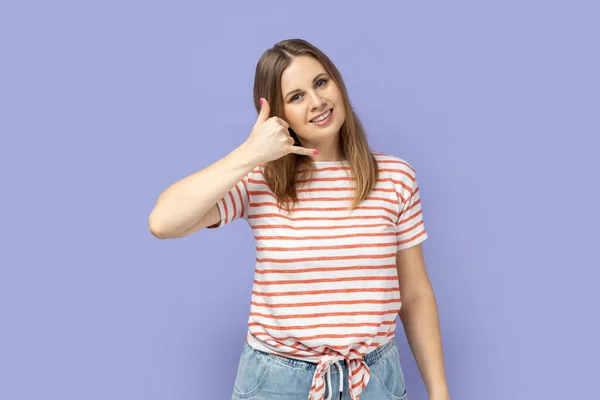 Hey handsome, call me. Blond woman wearing striped T-shirt flirting holding fingers near ear showing call gesture with toothy smile, answering call. Indoor studio shot isolated on purple background.