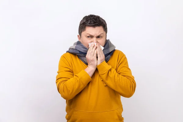穿着暖和围巾的男人在餐巾上打喷嚏 流鼻涕 季节性过敏 流感症状严重 头戴都市风格的帽衫 感觉不舒服 在白色背景下被隔离的室内拍摄 — 图库照片