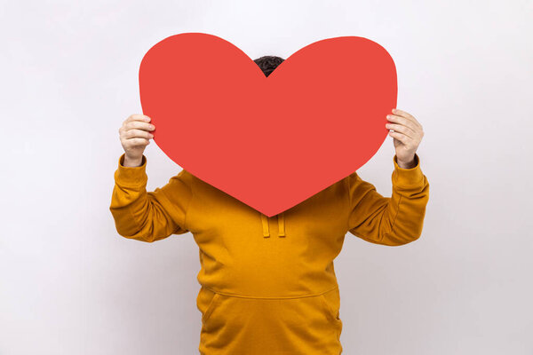 Unknown man hiding his face behind big red paper heart, making anonymous surprise, sending greeting card, wearing urban style hoodie. Indoor studio shot isolated on white background.