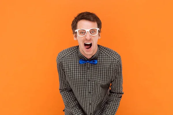 Crazy man nerd yelling loudly, opens mouth broadly, feels desperate after finding out about tragic events, wearing shirt with blue bow tie and glasses. Indoor studio shot isolated on orange background