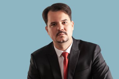 Kameraya bakan, dikkatli ve gülümsemeyen, kırmızı kravatlı siyah takım elbiseli, ciddi derecede konsantre olmuş bıyıklı bir adamın portresi. Açık mavi arkaplanda izole edilmiş kapalı stüdyo çekimi.