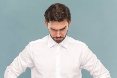 Bıyıklı, belinde kalça olan, alnının altından bakan, kızgın, açık mavi arka planda izole edilmiş kapalı mekan çekimleri giyen düşünceli, katı bir zorbanın portresi..