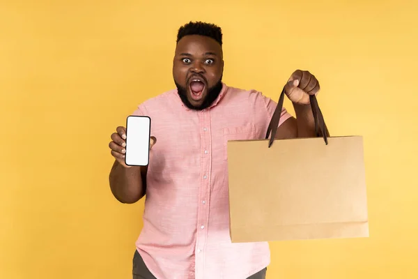 Portrait of shocked man in pink shirt holding shopping bag and cell phone with mock up, blank display for advertisement of online store mobile app. Indoor studio shot isolated on yellow background.