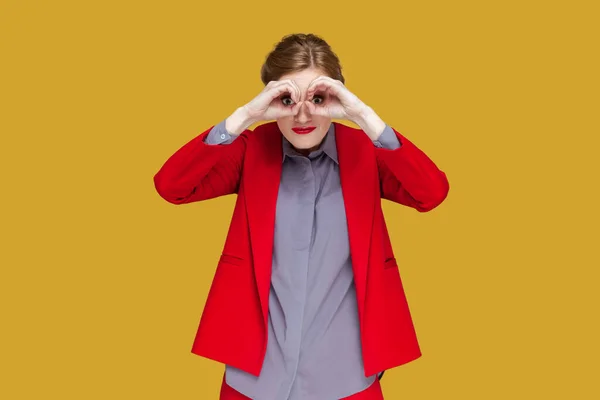 手で双眼鏡を作って立っている赤い唇を持つ驚きの集中女性の肖像画 遠くを見て 赤いジャケットを着て 黄色の背景に隔離された屋内スタジオショット — ストック写真