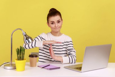 Çalışmaktan yorgun düşmüş genç bir kadın mola verme hareketi yapıyor, iş yerinde dizüstü bilgisayarla oturmak istiyor, profesyonel yorgunluk. Sarı arka planda kapalı stüdyo çekimleri.