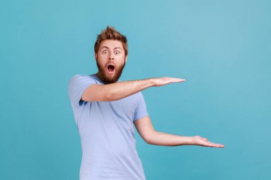 Genç yetişkin portresi şok olmuş sakallı adam reklam alanı sunuyor, kameraya bakıyor ağzı açık ve büyük gözleri var. Mavi arka planda kapalı stüdyo çekimi.