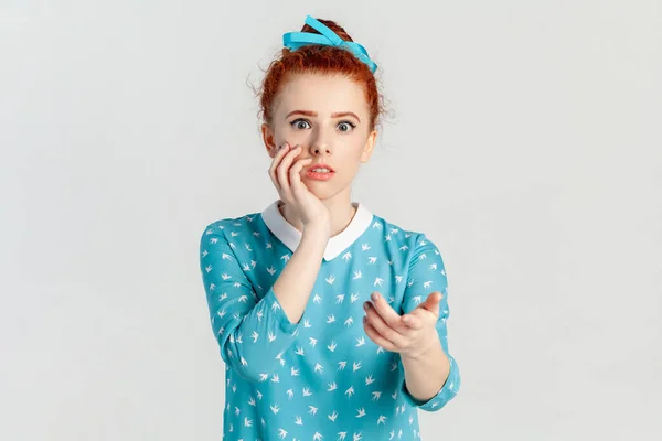Portrait of shocked surprised confused red haired woman with hair bun, pointing to camera with index finger, wearing blue dress. Indoor studio shot isolated on gray background.