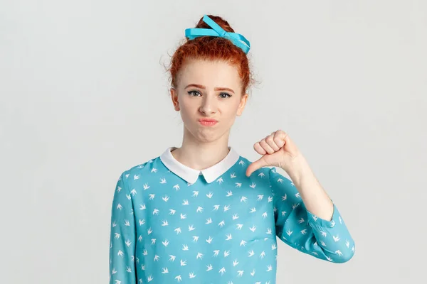 Portrait of unhappy sad upset red haired woman with hair bun, showing thumb down, being displeased, frowning her face, wearing blue dress. Indoor studio shot isolated on gray background.