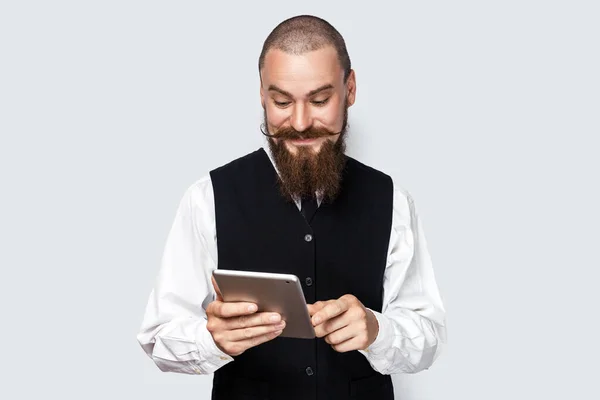 用平板电脑 与朋友聊天 在社交网络上阅读信息的面带笑容的 乐观的 英俊的 留着胡子和胡子的男人的画像 在灰色背景下被隔离的室内拍摄 — 图库照片