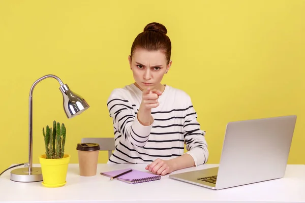 Irritated angry woman office worker in striped shirt pointing finger to camera, scolding and making you guilty, sitting at workplace in office. Indoor studio studio shot isolated on yellow background.