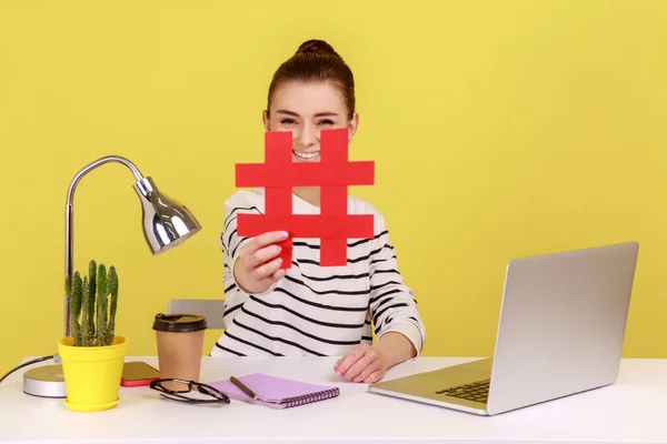 Happy delighted woman sitting at workplace with laptop and showing big red hashtag symbol, sharing viral content, tagged message. Indoor studio studio shot isolated on yellow background.