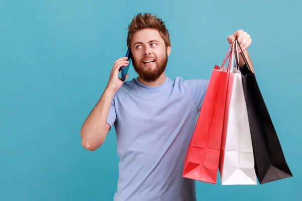Portrait of positive smiling bearded man holding shopping bags and talking on mobile phone, boasting her purchase, looking away. Indoor studio shot isolated on blue background.