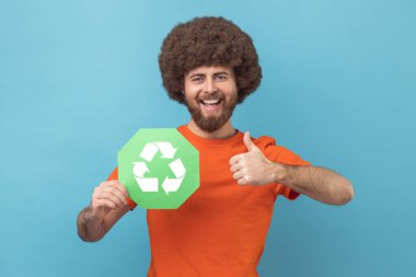 Turuncu tişört giyen, elinde atık geri dönüşüm sembolü tutan Afro saçlı bir adamın portresi baş parmağını gösteriyor, ekoloji için endişeleniyor. Mavi arka planda kapalı stüdyo çekimi.