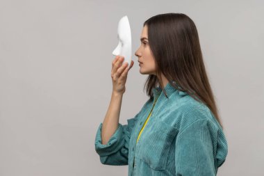 Beyaz maske takan, yüzünü kapatan, çoklu kişilik bozukluğu olan sıradan bir ceket giyen bir kadının yan görünümü. Gri arka planda izole edilmiş kapalı stüdyo görüntüleri.