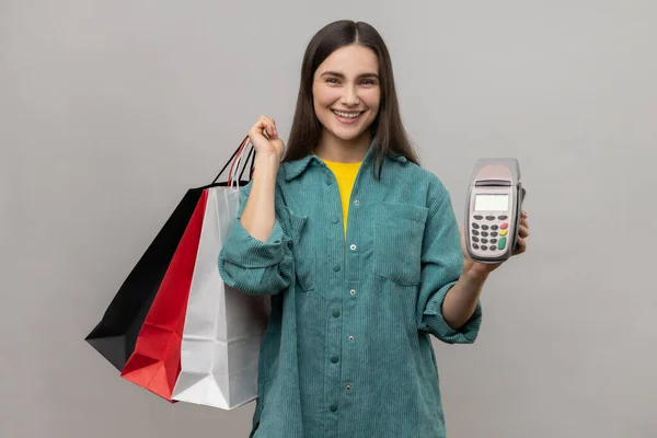 Positive smiling woman with dark hair holding payment terminal and paper shopping bags, easy express order and delivery, wearing casual style jacket. Indoor studio shot isolated on gray background.