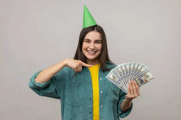 Lucky rich woman in party cone on head pointing at money, looking happiness by lottery win, celebrating big profit, wearing casual style jacket. Indoor studio shot isolated on gray background.