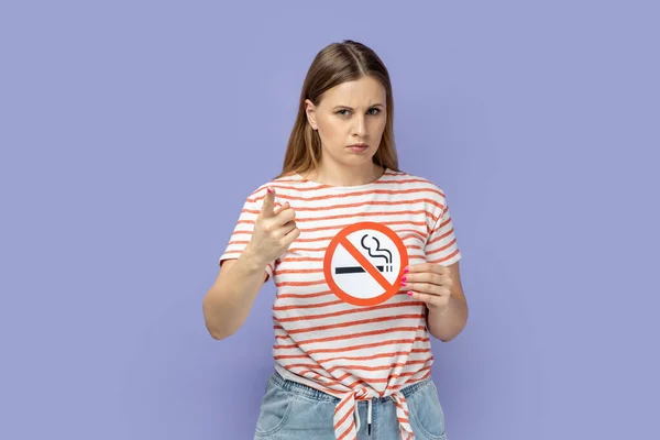 Portrait of strict bossy blond woman wearing striped T-shirt holding no smoking sign and pointing finger to camera, showing warning gesture. Indoor studio shot isolated on purple background.