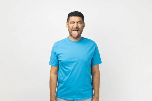 遊び心のあるハンサムなUnshaven男の肖像画青いTシャツを目を閉じて顔を眉をひそめて立って着て 彼の舌を示しています 灰色の背景に隔離された屋内スタジオショット — ストック写真