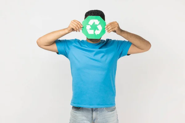 緑のリサイクル記号 ゴミの分類と環境保護の背後に隠れて顔を立って青いTシャツを着て男の肖像画は 緑を考えて 灰色の背景に隔離された屋内スタジオショット — ストック写真