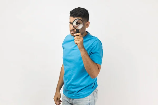 青いTシャツの立っているUnshaven男の肖像画は 拡大鏡を保持し 真正性を検証し 大きなズームの目でカメラを見て立っている 灰色の背景に隔離された屋内スタジオショット — ストック写真