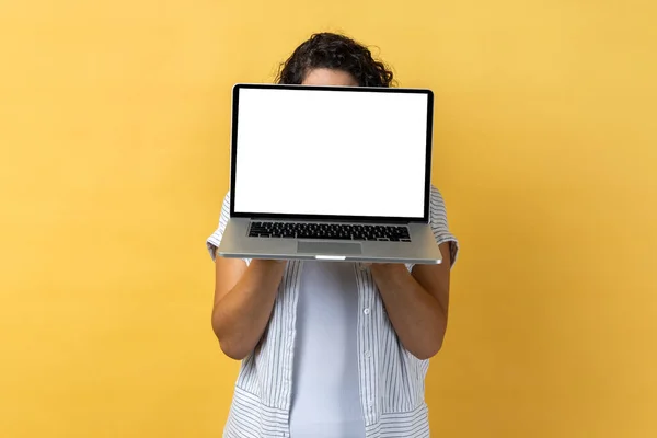 暗い波状の髪を空のディスプレイ 広告エリア プロモーションテキストのためのスペースとノートパソコンを保持している無名の女性の肖像画 黄色の背景に隔離された屋内スタジオショット — ストック写真