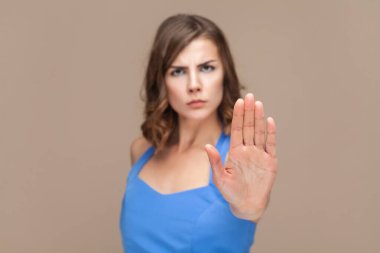 Kıvırcık saçlı, dur işareti sembolü gösteren, avuç içini kameraya çeken, mavi elbise giyen ciddi derecede katı bir kadının portresi. Açık kahverengi arka planda kapalı stüdyo çekimleri.