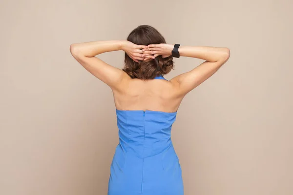 Portrait of anonymous woman with wavy hair standing backwards to camera with bare shoulders, keeps hands raised, wearing elegant blue dress. Indoor studio shot isolated on light brown background.