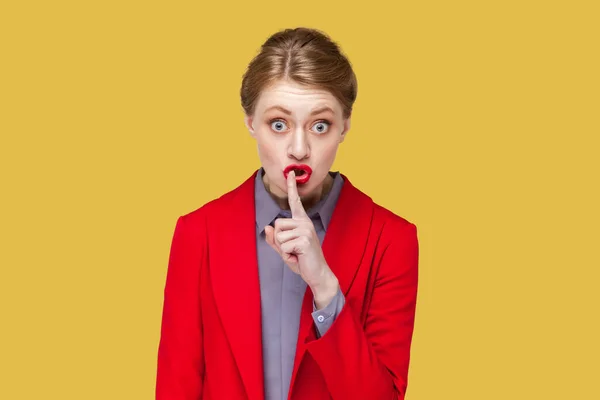 Portrait of strict bossy woman with red lips standing looking at camera, showing shh gesture, asking to be quiet, wearing red jacket. Indoor studio shot isolated on yellow background.