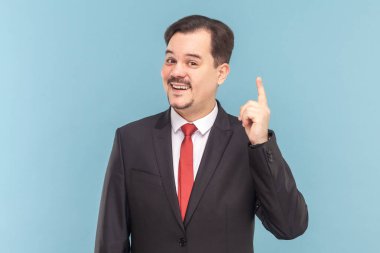Heyecanlı, bıyıklı, parmağını kaldırmış, ilginç bir fikri olan kırmızı kravatlı, siyah takım elbiseli bir adamın portresi. Açık mavi arkaplanda izole edilmiş kapalı stüdyo çekimi.