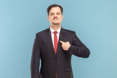 Bıyıklı gururlu, kendine güvenen, kendini gösteren, ciddi bir ifadeyle kameraya bakan, kırmızı kravatlı, siyah takım elbiseli bir adam. Açık mavi arkaplanda izole edilmiş kapalı stüdyo çekimi.