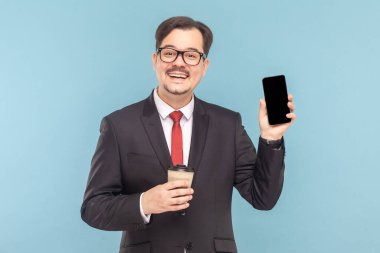 Bıyıklı gülümseyen adamın portresi cep telefonunu reklam alanına gösteriyor, kahve içiyor, kırmızı kravatlı siyah takım elbise giyiyor. Açık mavi arkaplanda izole edilmiş kapalı stüdyo çekimi.
