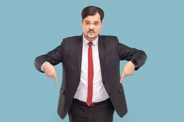 Here and right now. Portrait of serious angry man with mustache standing pointing finger down, wearing black suit with red tie. Indoor studio shot isolated on light blue background.