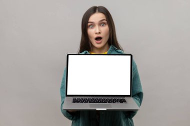 Terfi için beyaz beyaz ekranlı dizüstü bilgisayarı olan, ağzı açık kameraya bakan, günlük tarz ceket giyen bir kadın şaşırdı. Gri arka planda izole edilmiş kapalı stüdyo görüntüleri.