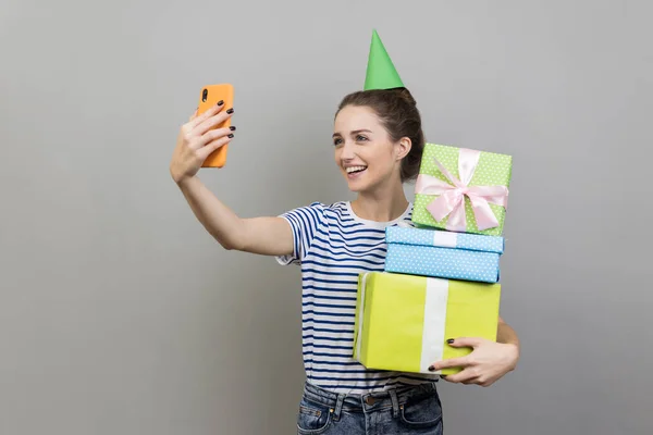 形象友好的微笑的女人穿着条纹T恤和派对锥 拿着一堆堆礼物盒 现场直播 在灰色背景下被隔离的室内拍摄 — 图库照片