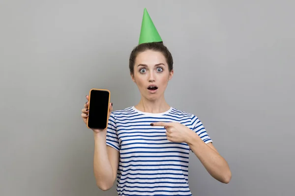 穿着条纹T恤和派对圆锥的惊讶女性的画像 显示了用空黑屏的手机做广告的情形 在灰色背景下被隔离的室内拍摄 — 图库照片