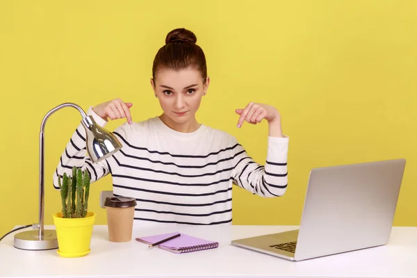 Here and right now. Young woman sitting at workplace with laptop and pointing fingers down, controlling and demanding deadline job finish. Indoor studio studio shot isolated on yellow background.