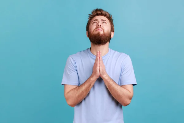 stock image Portrait of hopeless desperate bearded man folding hands in pray looking up, communicating with god, asking for forgiveness and blessing. Indoor studio shot isolated on blue background.